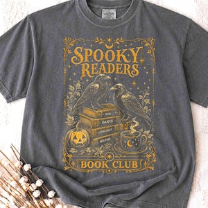 Pode incluir: Uma t-shirt cinzento-carvão de manga curta com uma impressão gráfica dourada. O design inclui dois corvos pousados numa pilha de livros com as etiquetas Poe, Dante, Lovecraft e Milton, além de uma abóbora, uma caneca e o texto Spooky Readers Book Club.