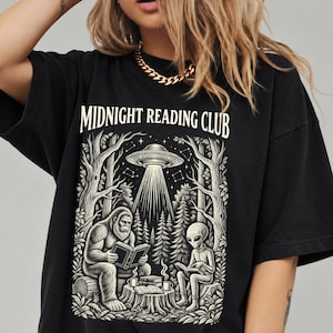 Op de afbeelding: Een zwart t-shirt met de tekst MIDNIGHT READING CLUB in een crèmekleurig lettertype. De afbeelding toont een Bigfoot en een alien die in een bos onder een UFO boeken lezen. De scène bevat bomen, een boomstronk en twee dampende mokken.