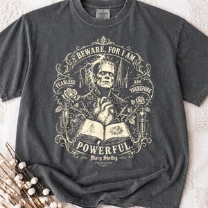 May include: A charcoal grey t-shirt featuring a cream-coloured graphic of Frankenstein's monster, an open book, roses, and laboratory equipment. The text reads: BEWARE, FOR I AM FEARLESS AND THEREFORE POWERFUL. Mary Shelley Frankenstein 1818.