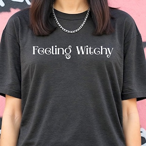 Op de afbeelding: Donkergrijs T-shirt met de witte tekst "Feeling Witchy". De top heeft een ronde hals en korte mouwen. Er wordt een zilveren ketting gedragen. De achtergrond is een roze en witte muur.