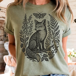 May include: Olive green t-shirt featuring a black illustration of a fox surrounded by botanical elements, including a butterfly, flowers, and mushrooms. The design is centered on the shirt, creating a nature-inspired aesthetic.