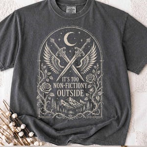 May include: A charcoal grey short-sleeve t-shirt featuring a cream-coloured graphic of two crossed swords with wings, a crescent moon, roses, and mountains. The text reads IT'S TOO NON-FICTIONY OUTSIDE, framed within an arched decorative border.