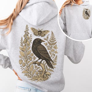 May include: A light gray hooded sweatshirt featuring a large back graphic of a black crow perched on a branch, surrounded by botanical illustrations of ferns, flowers, mushrooms, and a moth. A smaller version of the crow graphic is on the front.