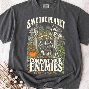 May include: A charcoal grey crew neck t-shirt featuring a graphic print with the text "SAVE THE PLANET" and "COMPOST YOUR ENEMIES." The design includes a compost bin, skulls, bones, a shovel, garden shears, mushrooms, and a pumpkin.