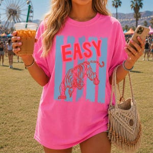 May include: A bright pink short-sleeved t-shirt featuring the word EASY in large red letters above a graphic of a tiger. The design is set against vertical light blue stripes. The shirt is worn with a beige macrame fringe bag with wooden beads.