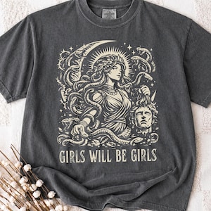 Puede incluir: Camiseta gris oscuro con un gráfico color crema de una mujer con serpientes como cabello, una luna creciente y estrellas. El texto "GIRLS WILL BE GIRLS" está impreso debajo. La camiseta tiene cuello redondo y mangas cortas.