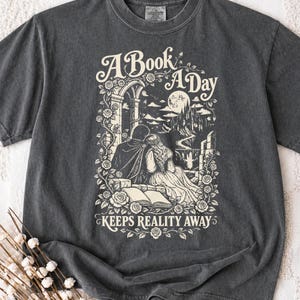 Może przedstawiać: Ciemnoszary t-shirt z kremowym nadrukiem graficznym przedstawiającym dwie postacie siedzące przy otwartej książce, zamku i różach. Tekst brzmi A Book A Day Keeps Reality Away, otoczony kwiatowymi pnączami i nocnym niebem z księżycem.