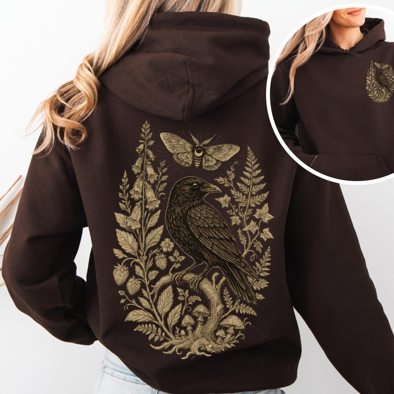 May include: A dark brown hooded sweatshirt featuring a large, detailed gold-toned graphic on the back depicting a raven perched on a branch, surrounded by ferns, mushrooms, flowers, and a moth. A smaller version of the graphic is on the front.