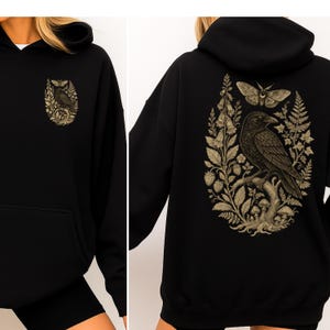 May include: A black hooded sweatshirt featuring a beige graphic print. The front displays a small illustration of a raven, moth, and foliage. The back features a larger, detailed print of a raven perched on a branch surrounded by moths, ferns, and mushrooms.