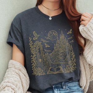 May include: A dark grey short-sleeved t-shirt featuring a gold-coloured line art graphic of a castle, a full moon, birds, and two figures standing before an open book, all framed by intricate floral vines and a stone archway design.