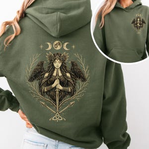 Op de afbeelding: Olijfgroene hoodie met een gedetailleerde illustratie op de rug. Het ontwerp toont een vrouw met lang haar, een kroon en een zwaard, geflankeerd door raven, met hemelse elementen erboven. Een kleinere versie van het ontwerp staat op de voorkant.
