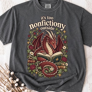 以下が含まれることがあります： チャコールグレーのTシャツ。開いた本を読む赤いドラゴンのグラフィックが描かれ、周囲には蔓、葉、バラがあしらわれています。ドラゴンのイラストの上部には、クリーム色のフォントで「it's too nonfiction outside」というテキストがプリントされています。