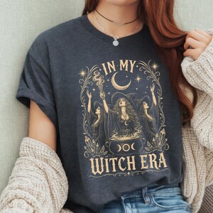 Op de afbeelding: Een donkergrijs t-shirt met een goudkleurige grafische print van drie figuren met heksenhoeden die rond een ketel staan. De tekst "IN MY WITCH ERA" is gedrukt in een decoratief lettertype, omringd door sterren, maansikkels en kristallen.