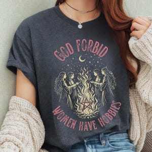 May include: Dark gray t-shirt with the text "GOD FORBID WOMEN HAVE HOBBIES" in pink. The graphic features four women around a bonfire with a pentagram, a crescent moon, and stars.