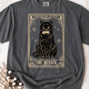 May include: A charcoal grey t-shirt featuring a graphic print of a black cat holding a mouse in its mouth, framed within a tarot card design labelled with the Roman numeral XV and the text THE MENACE in a cream-coloured decorative border.