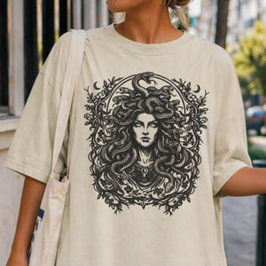 Op de afbeelding: Beige t-shirt met een zwart-wit illustratie van Medusa met slangen als haar, omlijst door een bloemen- en halve maan ontwerp. De afbeelding is gecentreerd op het shirt.
