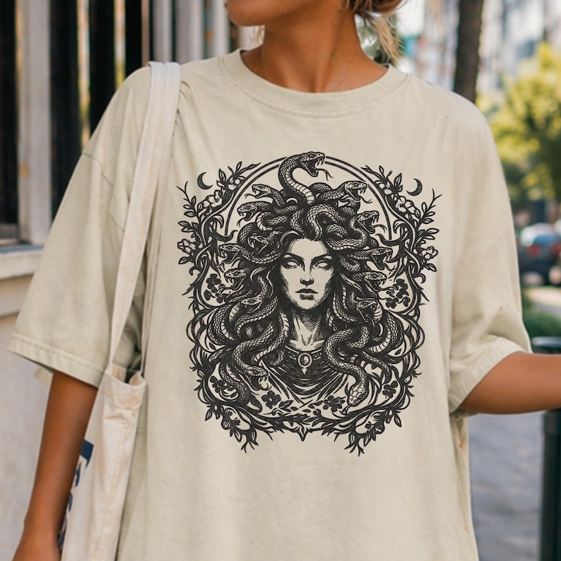 Op de afbeelding: Beige t-shirt met een zwart-wit illustratie van Medusa met slangen als haar, omlijst door een bloemen- en halve maan ontwerp. De afbeelding is gecentreerd op het shirt.
