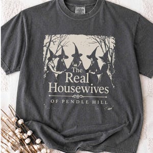 Könnte beinhalten: Ein anthrazitfarbenes Kurzarm-T-Shirt mit einem cremefarbenen Aufdruck von fünf Gestalten mit spitzen Hexenhüten zwischen Bäumen. Der Text lautet The Real Housewitches of Pendle Hill in einer Serifenschrift mit einer Zierlinie darunter.