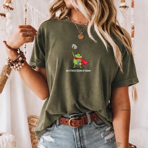 May include: An olive green short-sleeve t-shirt featuring a graphic of a green frog wearing a red cape, holding a mop, and carrying a cleaning bucket. The text below the frog reads WE STRESS CLEAN AT DAWN in white capital letters.