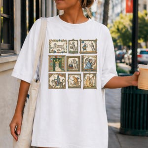 Op de afbeelding: Een wit T-shirt met korte mouwen voorzien van een raster van negen rechthoekige illustraties met antropomorfe katten in middeleeuwse sc&egrave;nes, waaronder een kat met een kroon, een kat die harp speelt en een kat die op een slak rijdt.