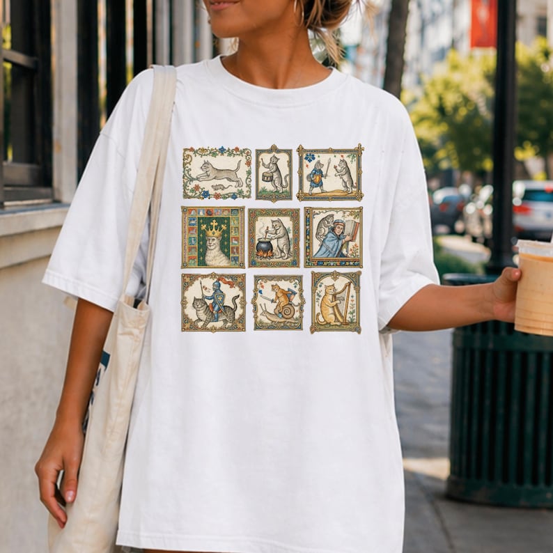 Op de afbeelding: Een wit T-shirt met korte mouwen voorzien van een raster van negen rechthoekige illustraties met antropomorfe katten in middeleeuwse sc&egrave;nes, waaronder een kat met een kroon, een kat die harp speelt en een kat die op een slak rijdt.