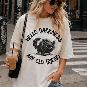 May include: A cream-coloured t-shirt with a black illustration of a grumpy cat. The text "Hello Darkness My Old Friend" is printed above and below the cat graphic in a bold, black font. The shirt has short sleeves and a relaxed fit.