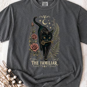 May include: A charcoal grey t-shirt featuring a graphic of a black cat holding a gold key in its mouth. The design includes pink roses, fern leaves, a crescent moon, stars, and the text THE FAMILIAR in a decorative gold font centred below the cat.