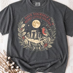 Może przedstawiać: Ciemnoszary t-shirt z krótkim rękawem z nadrukiem przedstawiającym kamienny monument, ptaka i pełnię księżyca w wieńcu z ostrokrzewu. Napis głosi "HAVE YOURSELF A MERRY LITTLE SOLSTICE", a na dole znajduje się symbol potrójnego księżyca.