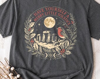 May include: A charcoal gray short-sleeve t-shirt featuring a graphic print of a stone monument, a bird, and a full moon surrounded by a holly wreath. The text reads, "HAVE YOURSELF A MERRY LITTLE SOLSTICE," with a triple moon symbol at the bottom.