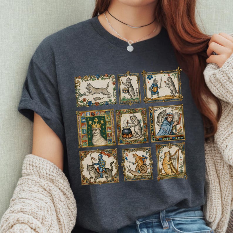 Op de afbeelding: Een donkergrijs t-shirt met een raster van negen rechthoekige illustraties met katten in middeleeuwse sc&egrave;nes, waaronder een kat met een kroon, een kat die harp speelt, een kat op een slak en katten die verschillende activiteiten uitvoeren.