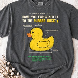 May include: A charcoal gray t-shirt featuring a yellow rubber duck graphic with technical diagram lines and labels. The text reads: HAVE YOU EXPLAINED IT TO THE RUBBER DUCK? and includes a code snippet: def debug(problem, duck): explain(problem, duck) return "oh."