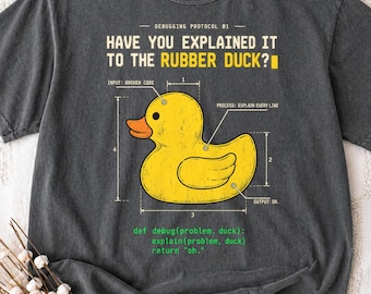 May include: A charcoal gray t-shirt featuring a yellow rubber duck graphic with technical diagram lines and labels. The text reads: HAVE YOU EXPLAINED IT TO THE RUBBER DUCK? and includes a code snippet: def debug(problem, duck): explain(problem, duck) return "oh."