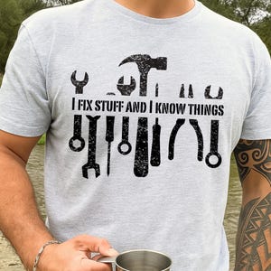 Pode incluir: Camiseta cinza claro com um gráfico preto de ferramentas e o texto "I FIX STUFF AND I KNOW THINGS". As ferramentas incluem um martelo, uma chave inglesa e uma chave de fenda. A camiseta é feita de um material macio.
