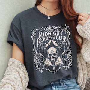 May include: A dark charcoal grey t-shirt featuring a white graphic print of a skull, two ravens, and an open book with the text "MIDNIGHT READERS CLUB" at the top and "Read Late. Stay Cursed." printed on the pages of the open book.