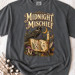 Puede incluir: Una camiseta gris carbón con un estampado gráfico que incluye el texto "MIDNIGHT MISCHIEF" sobre un cuervo negro posado en un libro abierto. El diseño muestra una luna creciente, una vela encendida, una pluma, tinta, setas y motivos botánicos.