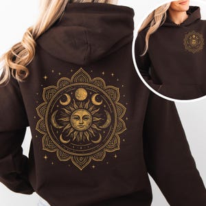 Puede incluir: Una sudadera con capucha marr&oacute;n oscuro con un dise&ntilde;o dorado de sol y luna. La parte trasera presenta un mandala detallado con un sol y una luna, y una versi&oacute;n m&aacute;s peque&ntilde;a en el pecho. El dise&ntilde;o tiene un tema celestial.