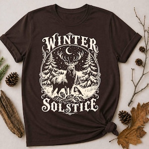 以下が含まれることがあります： ダークブラウンの半袖Tシャツ。雪の積もった森に立つ鹿、松の木、三日月、小さな鳥がクリーム色のグラフィックでプリントされています。装飾的なフォントで「WINTER SOLSTICE」という文字がデザインされています。