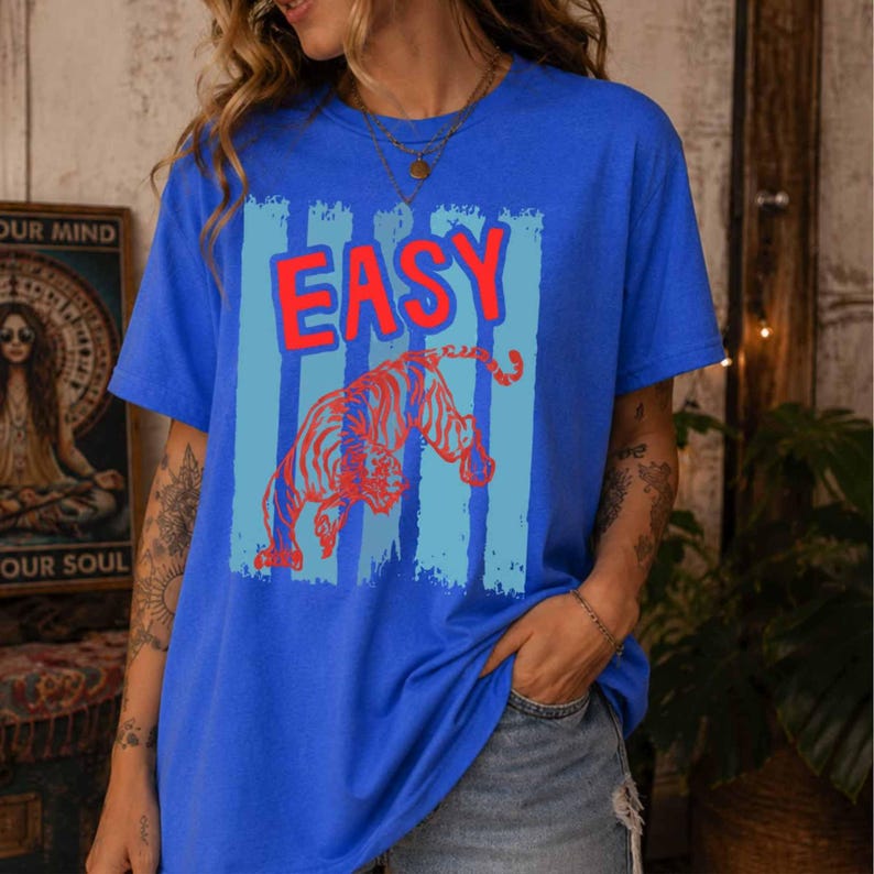 May include: A bright blue short-sleeve t-shirt featuring a graphic print of a red tiger and the word "EASY" in red letters. The design is set against a background of vertical light blue stripes. The shirt has a crew neckline and a relaxed fit.