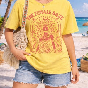 Pu&ograve; includere: T-shirt giallo con la scritta "THE FEMALE GAZE" e un'illustrazione rossa di una donna con serpenti al posto dei capelli. La maglietta &egrave; abbinata a pantaloncini di jeans e una borsa a tracolla fantasia.