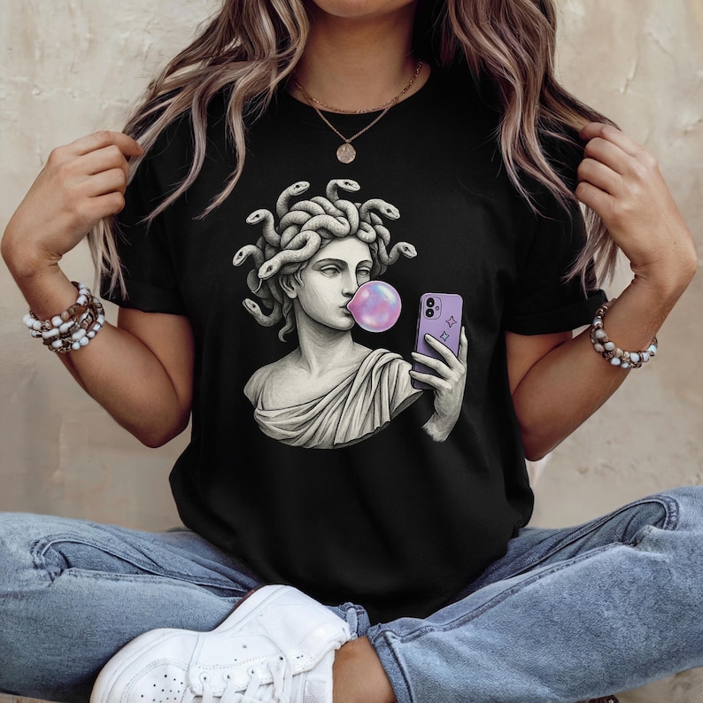 May include: Black t-shirt with a graphic of Medusa blowing a bubblegum bubble and taking a selfie with a purple phone. The Medusa illustration is in a classical statue style.