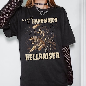 Może przedstawiać: Czarny t-shirt z grafiką przedstawiającą kobietę jadącą na tygrysie, trzymającą pochodnię i miecz. Tekst na koszulce brzmi "In a world of HANDMAIDS Be a HELLRAISER" w postarzanym kroju pisma. Projekt jest w odcieniach brązu i złota.