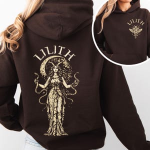 May include: A dark brown hoodie featuring a gold illustration of Lilith, a woman with horns and snakes, on the back. The word "LILITH" is arched above the illustration. The front of the hoodie has the word "LILITH" and a smaller illustration.