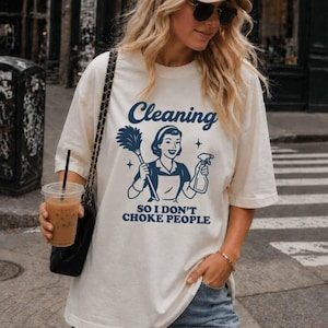 May include: Cream-colored t-shirt with a blue graphic that reads "Cleaning SO I DON'T CHOKE PEOPLE." The graphic features a vintage-style illustration of a woman holding a feather duster and a spray bottle. The shirt has short sleeves and a relaxed fit.