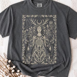 May include: A charcoal grey short-sleeve t-shirt featuring a cream-coloured graphic print of a central figure with antlers, flanked by two deer, an owl, and a bird. The design includes a labyrinth symbol, mushrooms, and an oak leaf border frame.