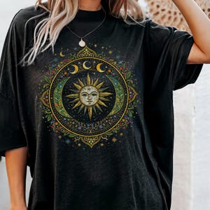 May include: Black t-shirt featuring a celestial design with a sun face, crescent moons, and stars. The design is circular, with intricate floral and dot details in shades of green, gold, and blue.