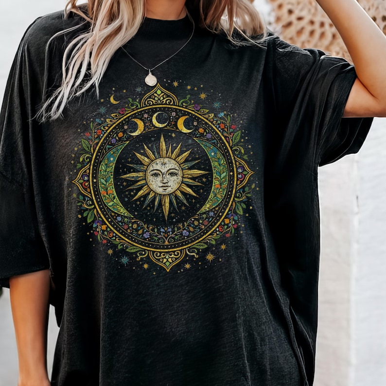 May include: Black t-shirt featuring a celestial design with a sun face, crescent moons, and stars. The design is circular, with intricate floral and dot details in shades of green, gold, and blue.