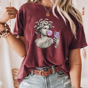 May include: Burgundy t-shirt with a graphic of a Medusa bust blowing a bubblegum bubble while taking a selfie with a purple phone. The Medusa bust is in grayscale, with snakes for hair. The shirt has a relaxed fit.