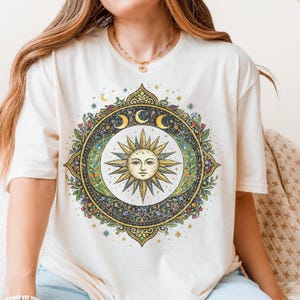 May include: Cream-colored t-shirt featuring a detailed circular design with a sun face at the center, surrounded by crescent moons, stars, and floral elements. The design is in shades of yellow, green, and blue.