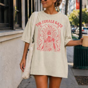 Pu&ograve; includere: T-shirt oversize color crema con grafica rossa di Medusa e la scritta "THE FEMALE GAZE". La grafica presenta una donna con serpenti al posto dei capelli, circondata da elementi decorativi. La maglietta &egrave; in stile casual e confortevole.
