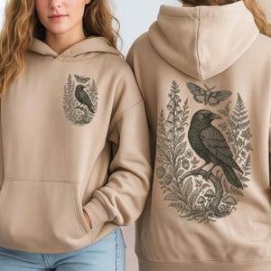 May include: A beige hooded sweatshirt featuring a black ink-style illustration of a raven perched on a branch, surrounded by ferns, mushrooms, and a moth. The front chest displays a smaller version of the same raven and moth graphic design.